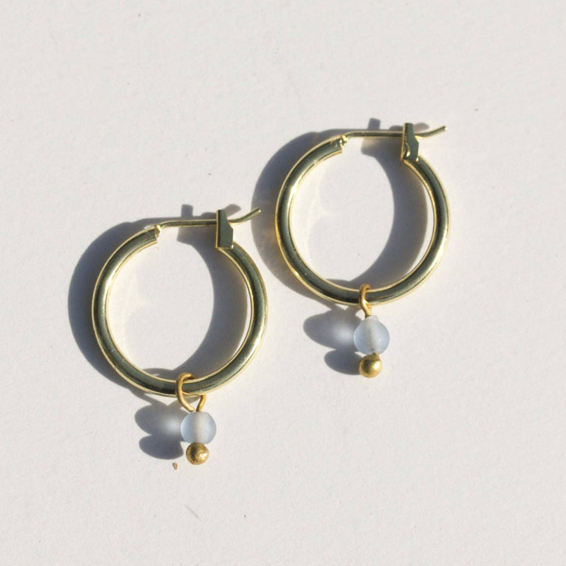 Two gold brass huggie hoop earrings with a small blue bead of sea glass hanging from the bottom.