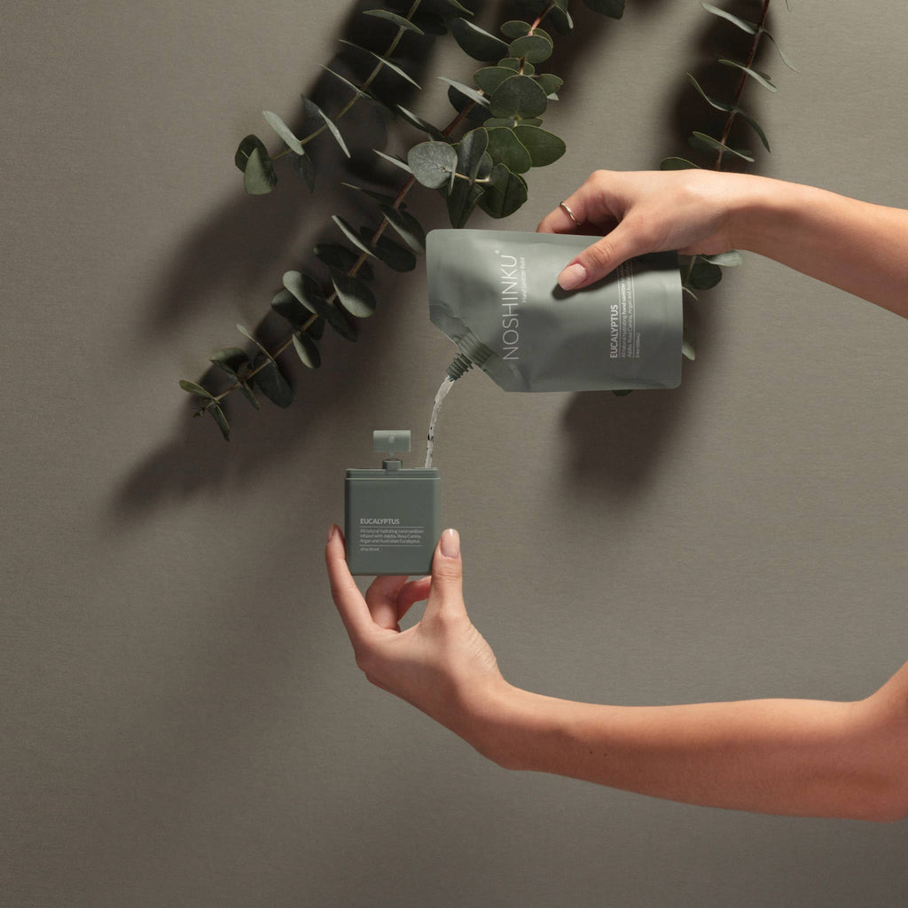 A pair of hands pouring the Eucalyptus Refill Pouch into a Sanitizer Sprayer in front of a green wall and inverted branches of eucalyptus.