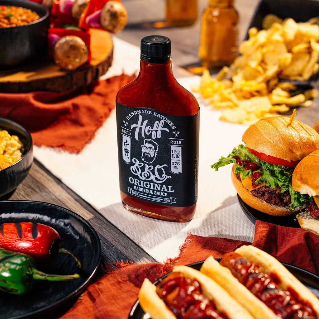 Hoff's Orinal BBQ Sauce surrounded by burgers, hot dogs, chips, peppers, and other bbq foods.