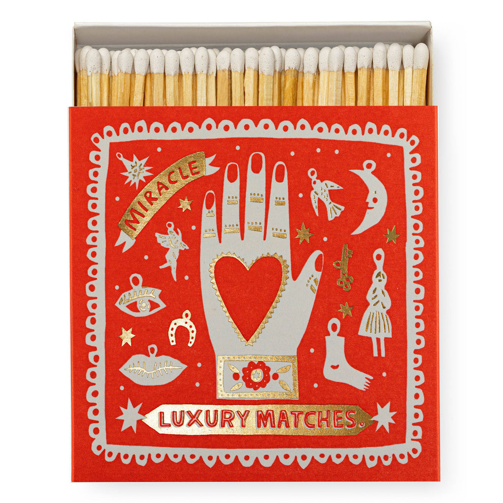 A red and gold foiled square matchbox illustrated with a heart in a hand surrounded by charms of body parts.