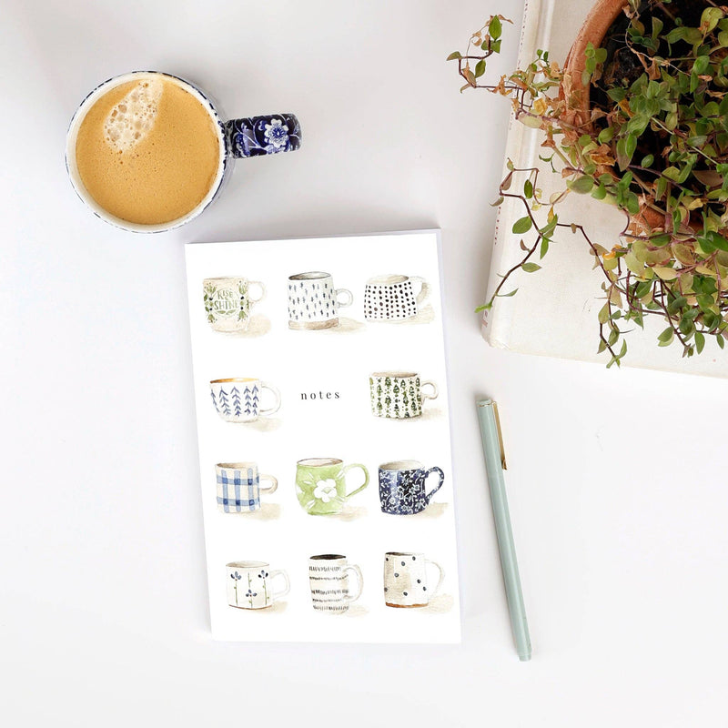 The Mugs Notebook resting on a desk next to a potted plant and a latte in a mug.