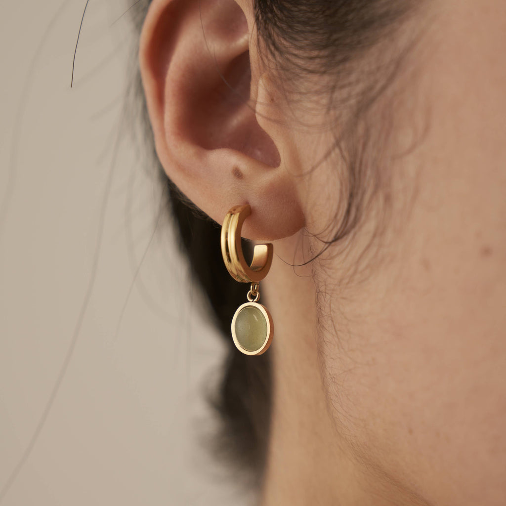 A person wearing a gold plated stainless steel huggie hoop with a green aventurine stone encased in gold hanging from the bottom.