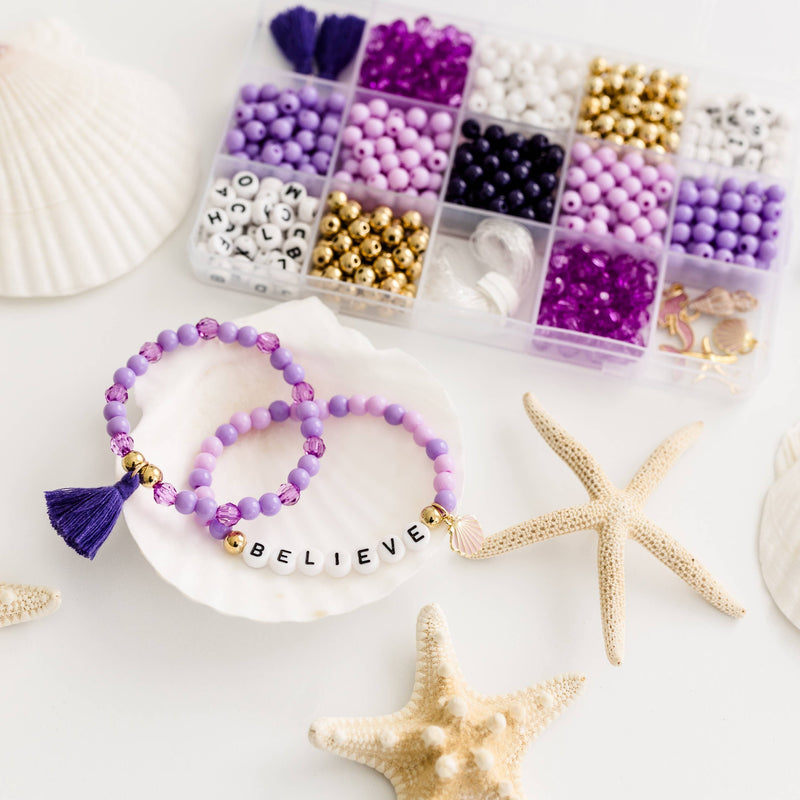 Two purple beaded bracelets decorated with the word "Believe" and charms surrounded by the Mermaid at heart Bracelet Kit, starfish, and shells.