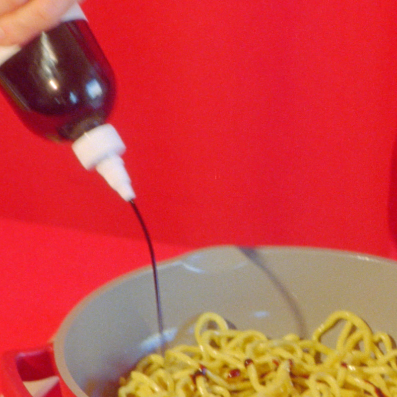 A person squeezing Woon Stir Fry Sauce onto a bowl of noodles.