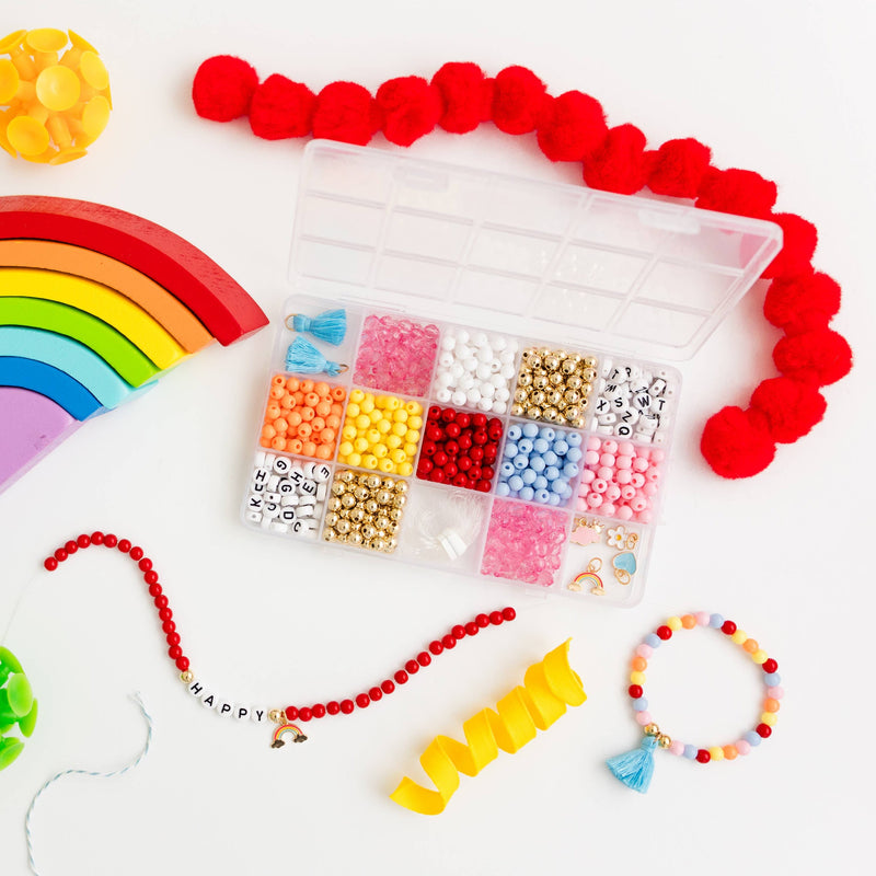 An open storage box of colorful beads and charms next to two finished bracelets, pompoms, and other colorful items.