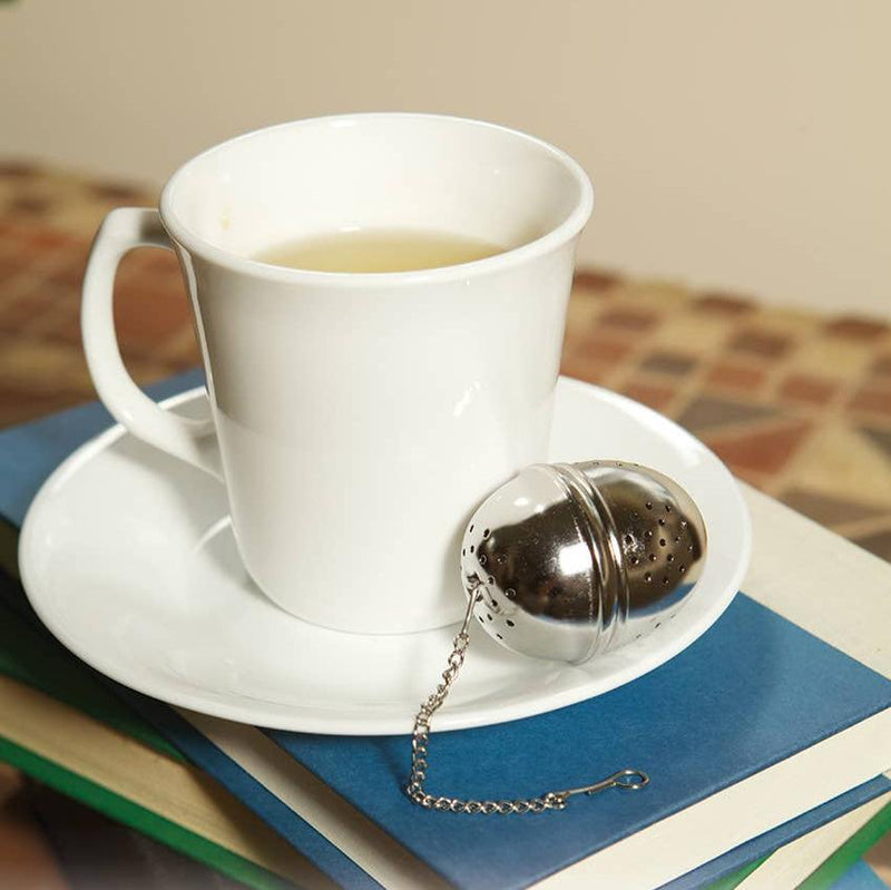 A tea ball resting on a tea cup saucer next to a cup of green tea.