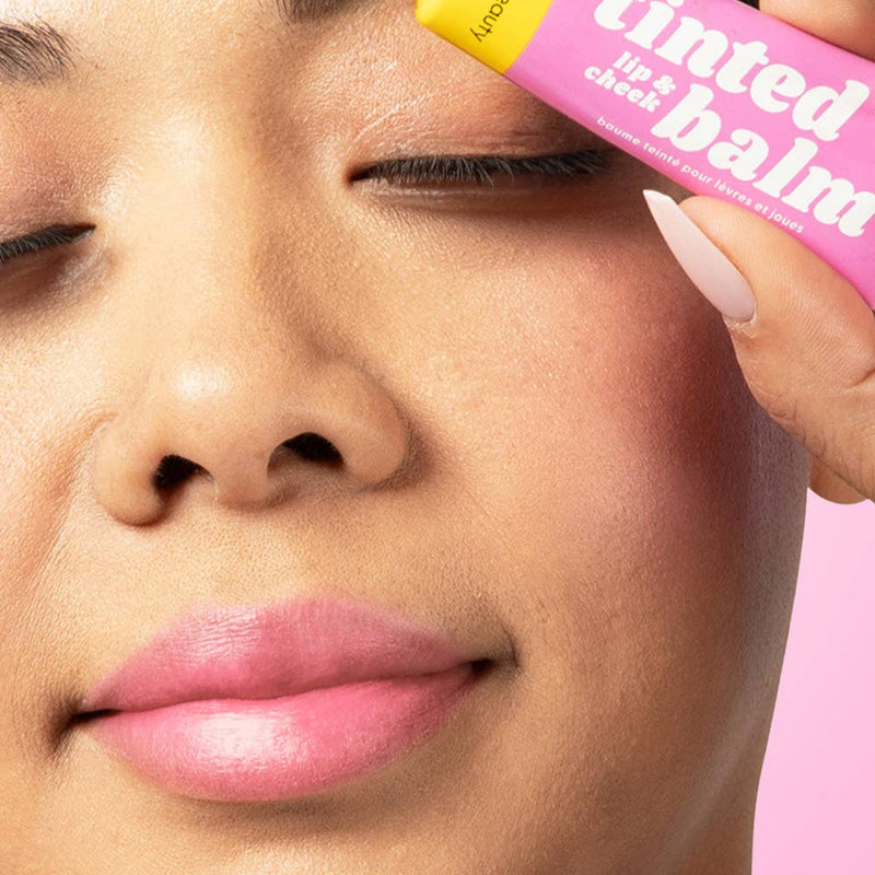 A person with medium tone skin wearing Pink Me Up on their eyelids, cheekbones, and lips while holding up the tube next to their eye.