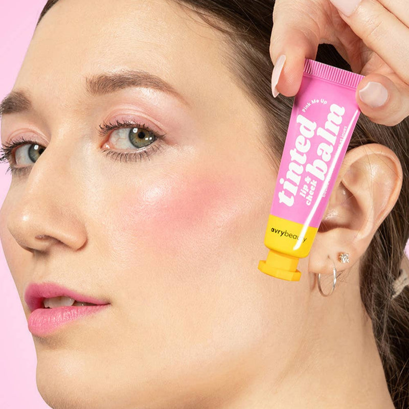 A person with light tone skin wearing Pink Me Up on their eyelids, cheekbones, and lips while holding the tube next to their face.