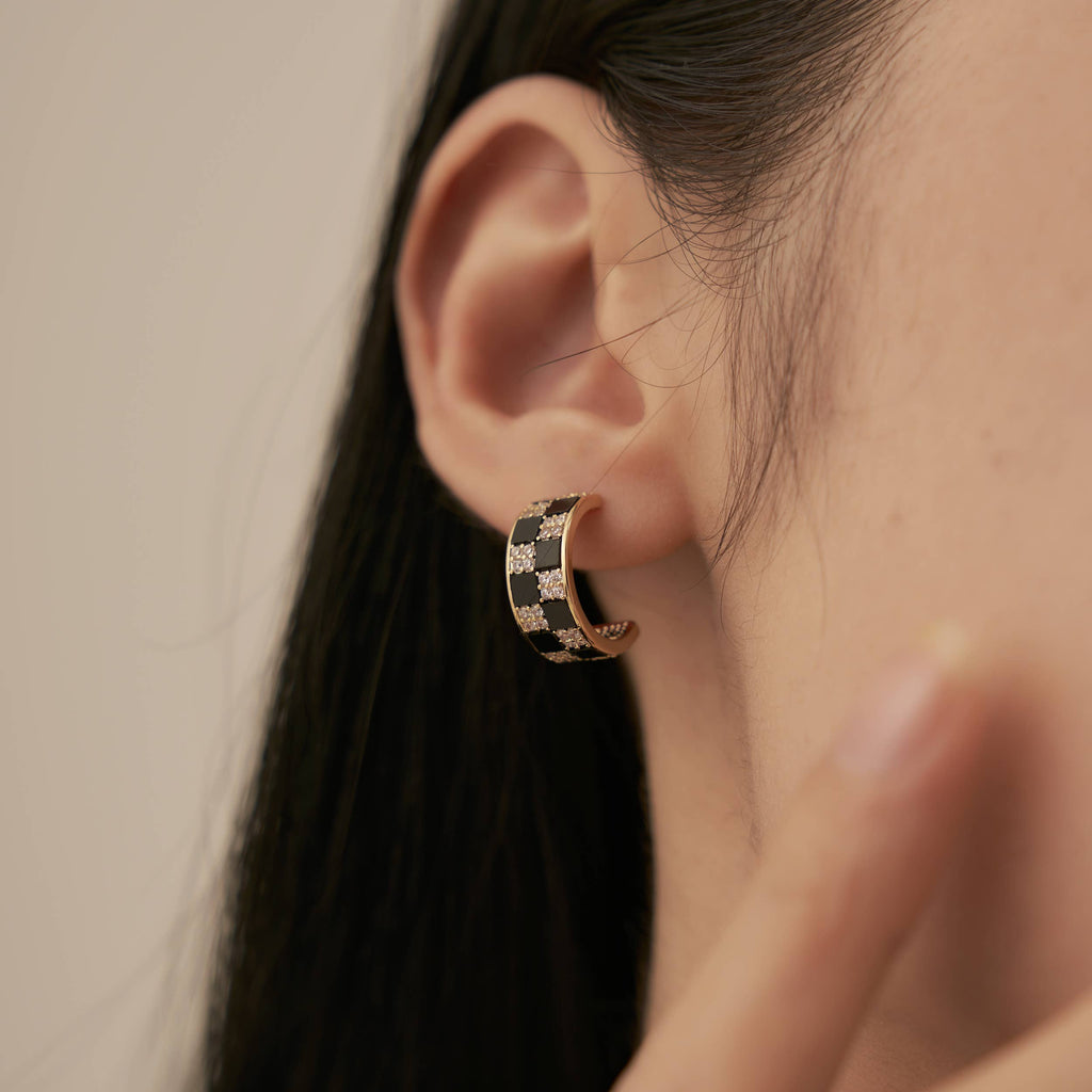 A person wearing a gold hoop earring inlaid with white and black gems in a checkerboard pattern.