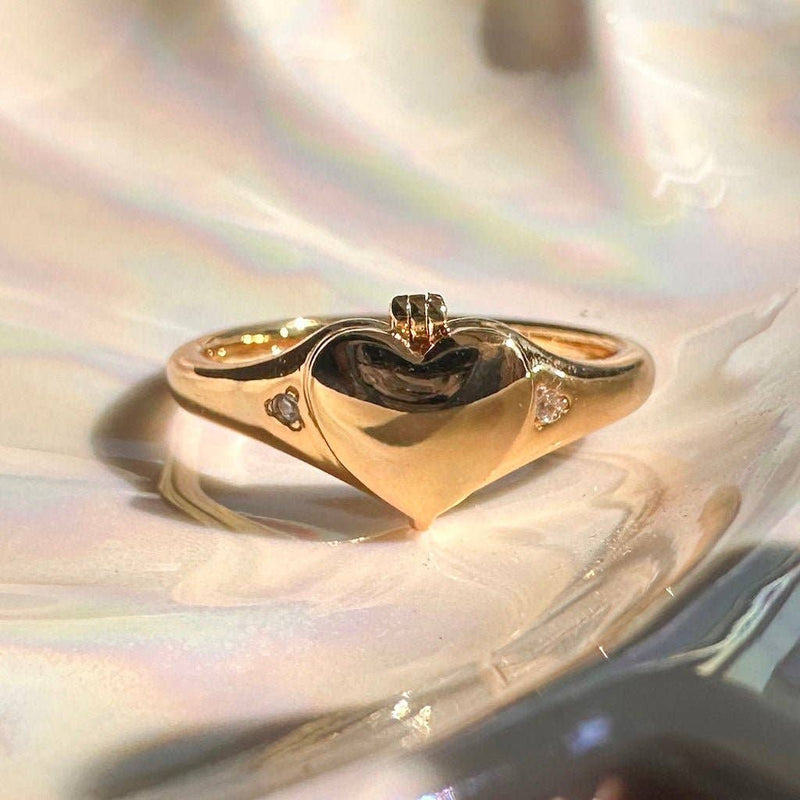 A gold plated ring with a heart locket face with two tiny embedded crystals on either side.