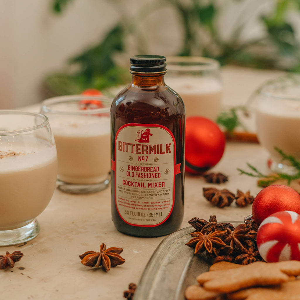 A bottle of No.7 Gingerbread Old Fashioned Mixer surrounded by glasses of No.7 Nog, ornaments, cedar, gingerbread cookies, and star anise.