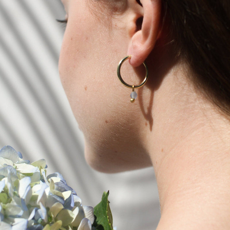 A person wearing the Sea Glass Huggies in Blue Sky next to a bouquet of hydrangeas in a similar color.