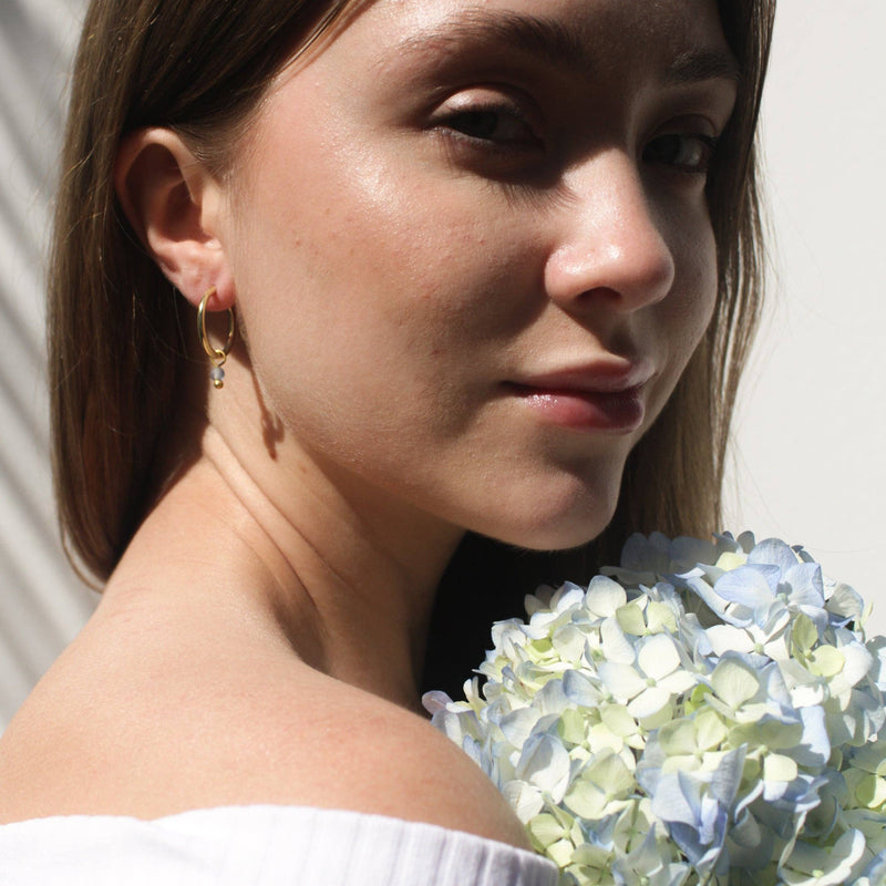  A person wearing the Sea Glass Huggies in Blue Sky next to a bouquet of hydrangeas in a similar color.