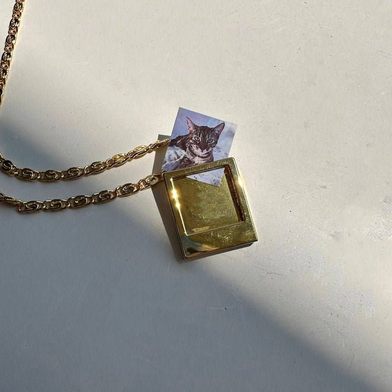 A gold plated polaroid frame charm with a tiny photo of a cat sliding into its photo slot.