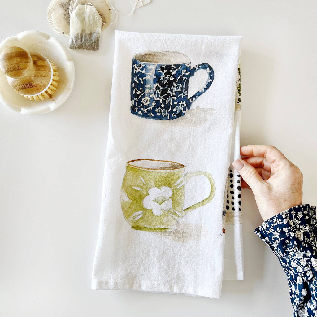 A folded Mugs Tea Towel illustrated with watercolor mugs being held by a hand next to a dish brush and a pile of teabags.