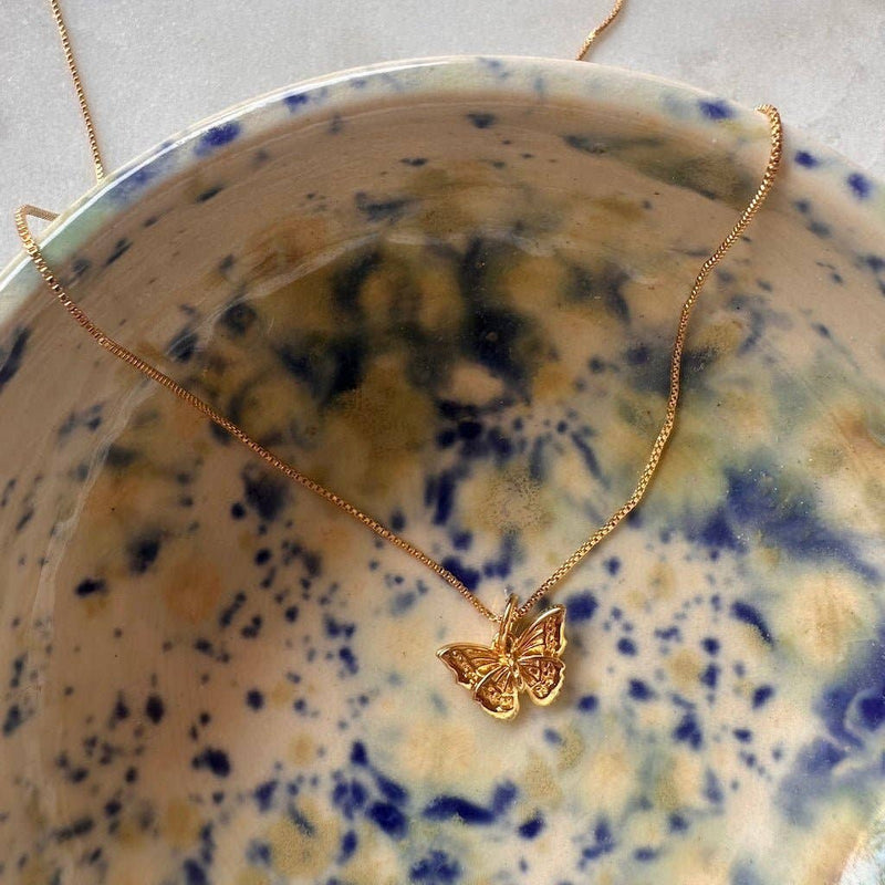 A Butterfly Necklace in Gold draped over a blue and cream speckled bowl.