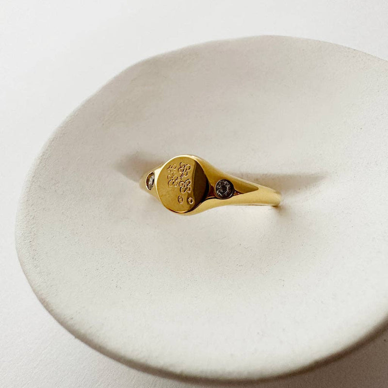 The Flora Signet Ring in Gold positioned upwards on a ceramic plate.