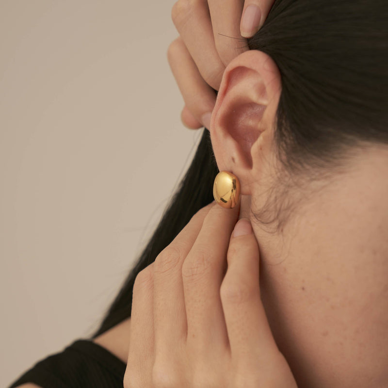 A person wearing a glossy finish gold plated stainless steel stud earring in the shape of a round rectangle.