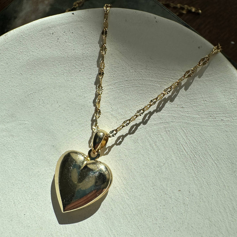 The Golden Heart Locket necklace resting on a ceramic plate in the sun.