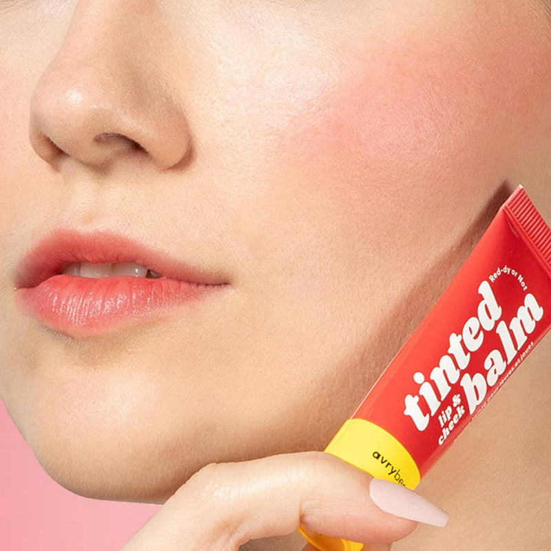 A person with light tone skin wearing Red-dy or Not Lip & Cheek Tinted Balm on their cheekbones and lips while holding up the tube next to their jaw.