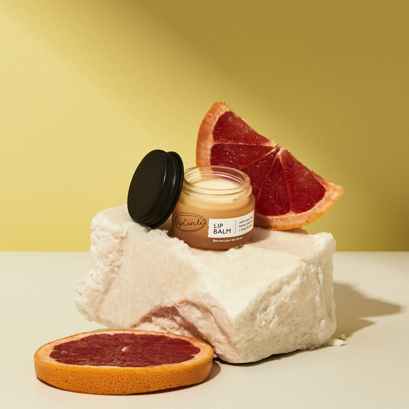 An open jar of Lip Balm resting on a stone surrounded by slices of grapefruit.