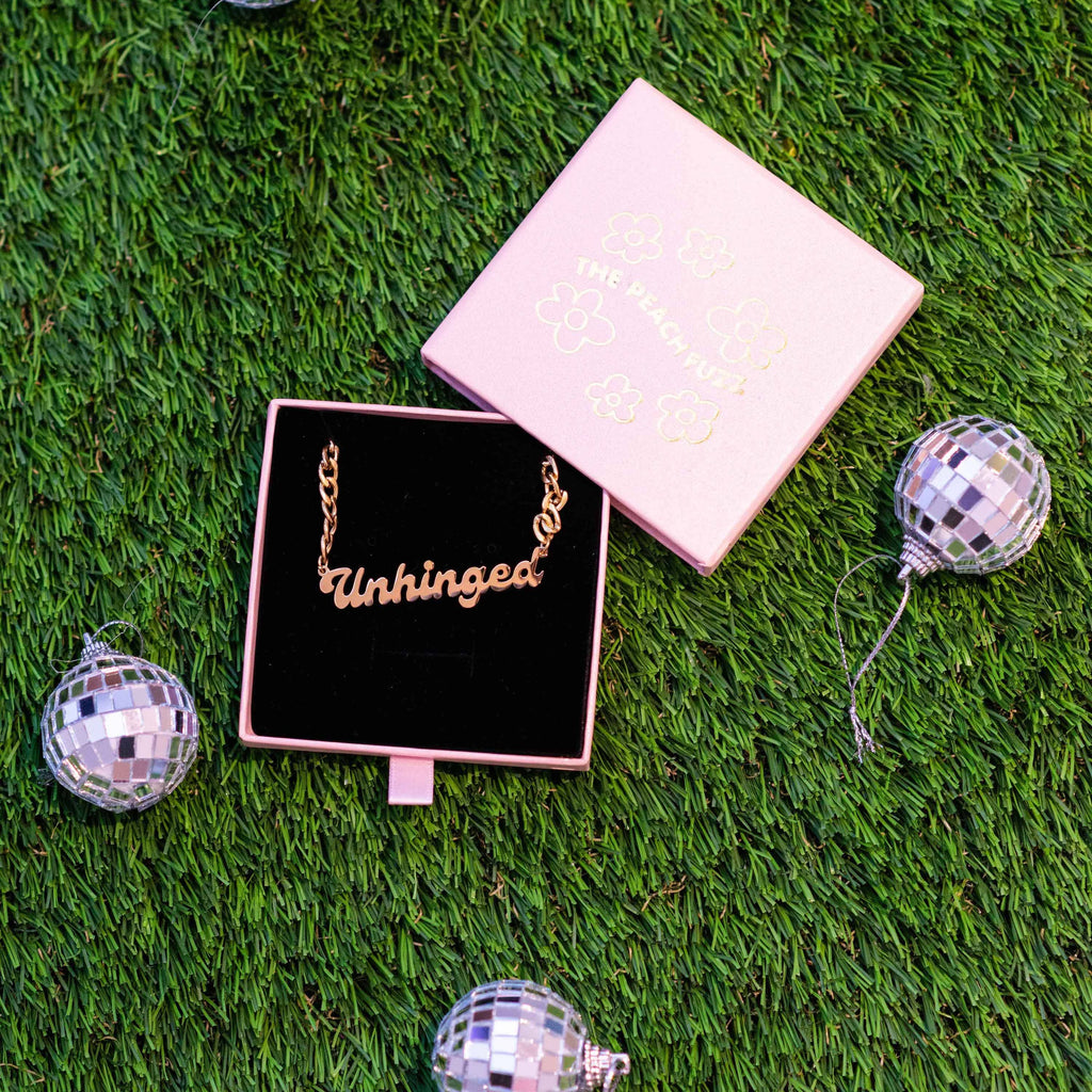 A gold chain necklace with a plate that says "Unhinged" in a pink and black jewelry box.