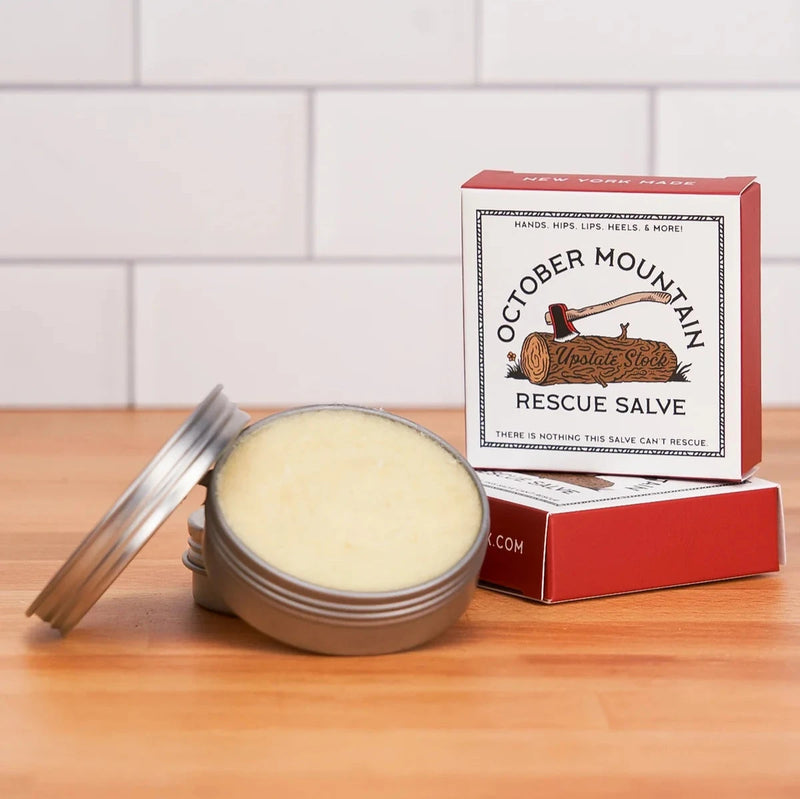 An open tin of October Mountain Rescue Salve next to its packaging stacked on another salve.