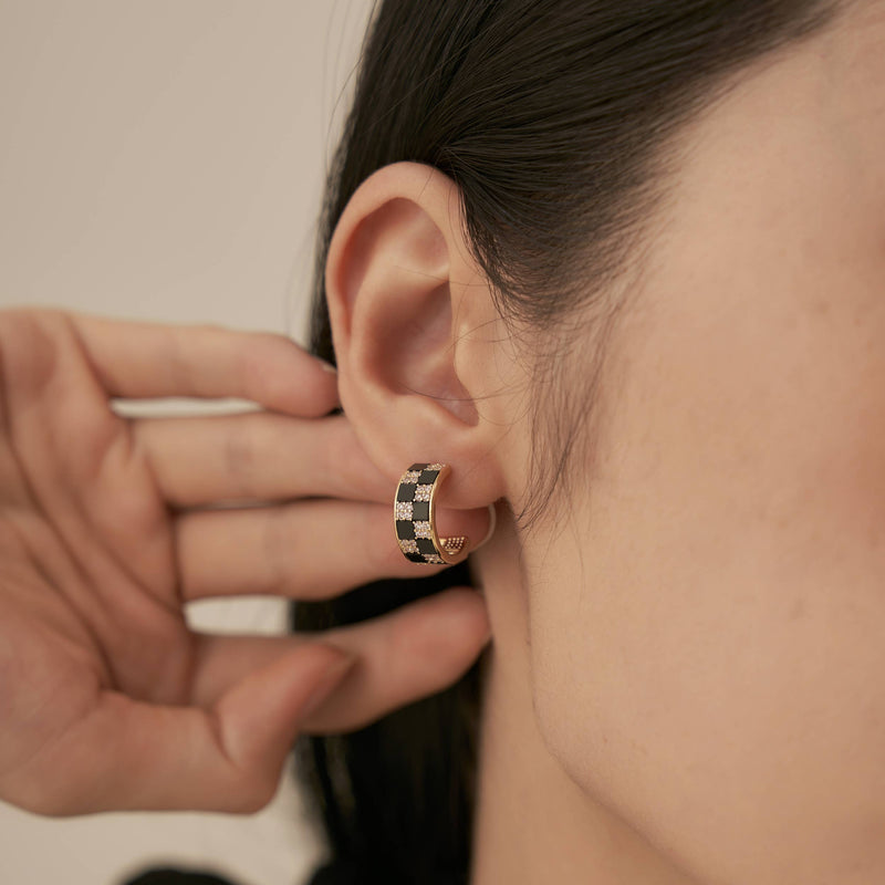 A person wearing a gold hoop earring inlaid with white and black gems in a checkerboard pattern.