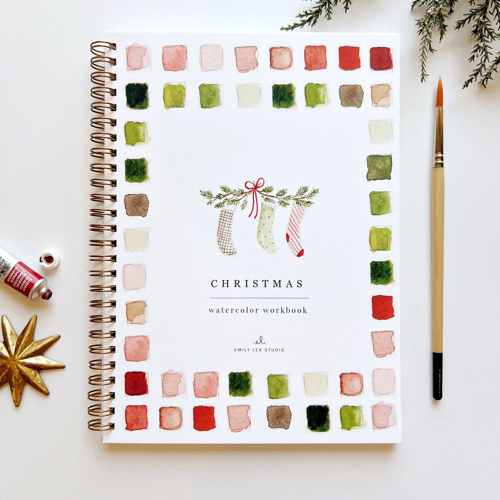 A Christmas Watercolor workbook with a watercolor illustration of three green and red stockings hanging on pine branches surrounded by green, pink, and red watercolor swatches.