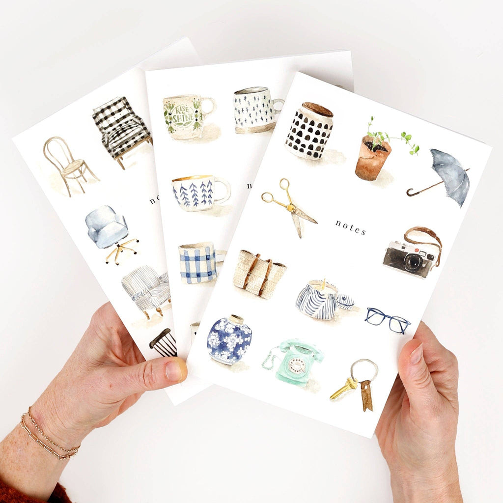 A person fanning out three notebooks, each illustrated with different watercolor objects.