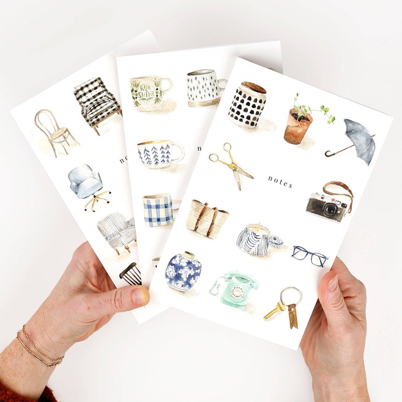 A person fanning out three notebooks, each illustrated with different watercolor objects.