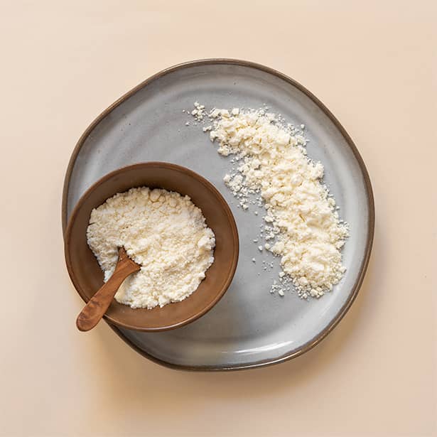 A bowl of white cheddar cheese powder with a wooden spoon resting on a ceramic plate with more white cheddar cheese powder.