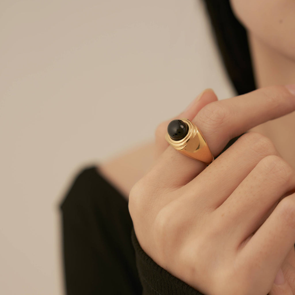 A person wearing a gold plated stainless steel ring with a black onyx stone embedded in a square encasement.