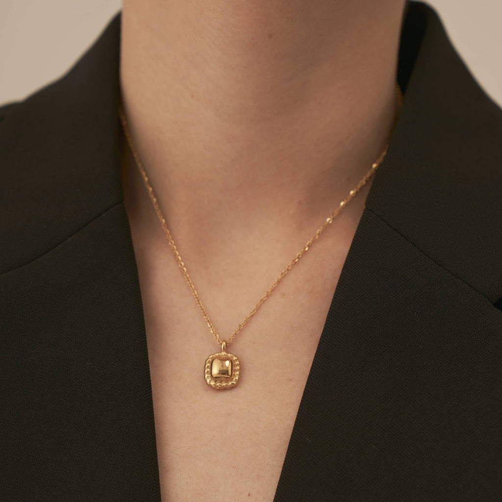 A gold necklace chain with a square pendant with braided ornamentation around it.