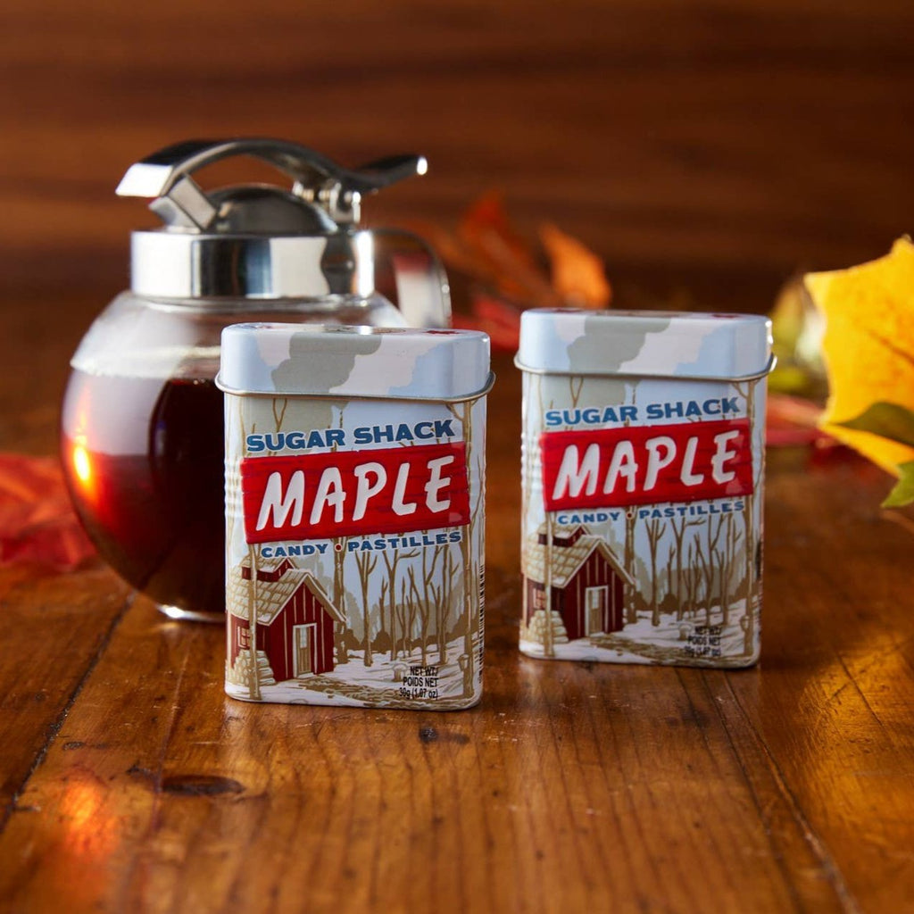 Two Sugar Shack Maple Candy Tins in front of a jar of maple syrup and autumn leaves.