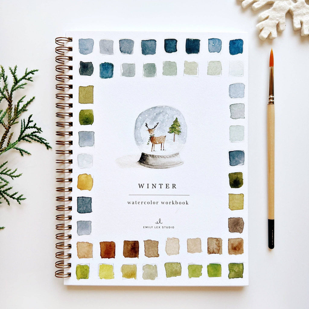 A Winter Watercolor Workbook with a watercolor illustration of a deer and a tree in a snow globe surrounded by watercolor paint swatches.