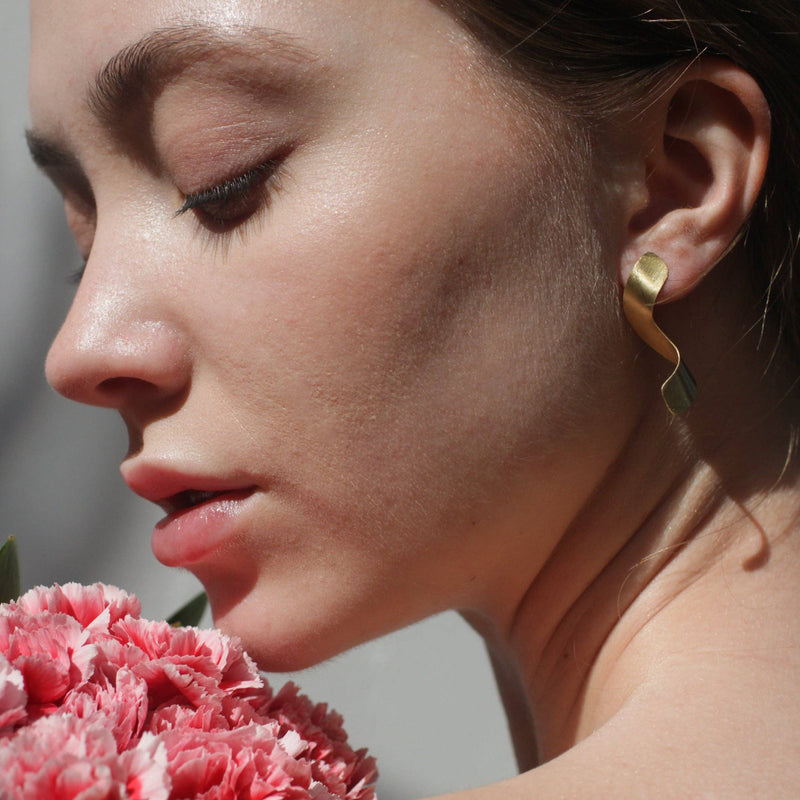 A person wearing the Golden Noodle Earrings while holding a bouquet of pink flowers to their face.