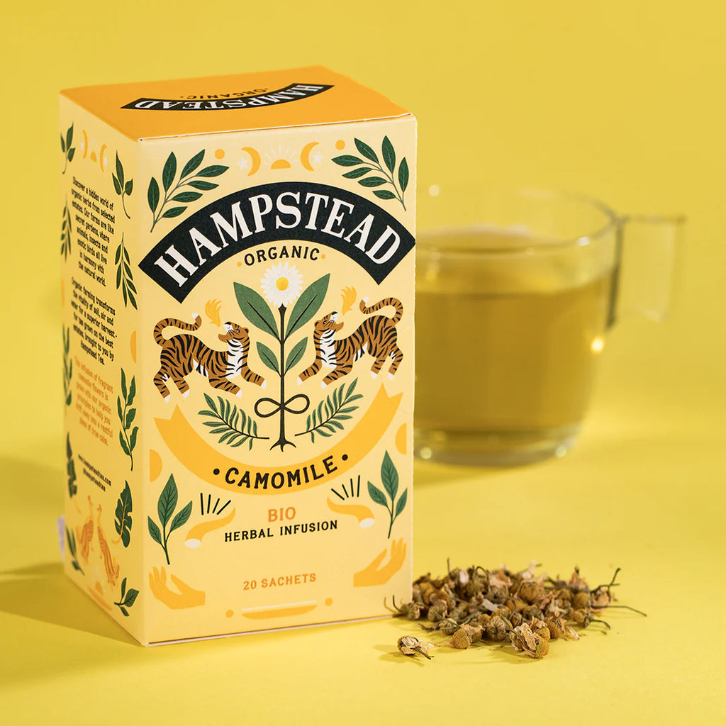 A box of Hampstead Camomile Tea next to a pile of dried chamomile flowers and golden chamomile tea in a glass cup.