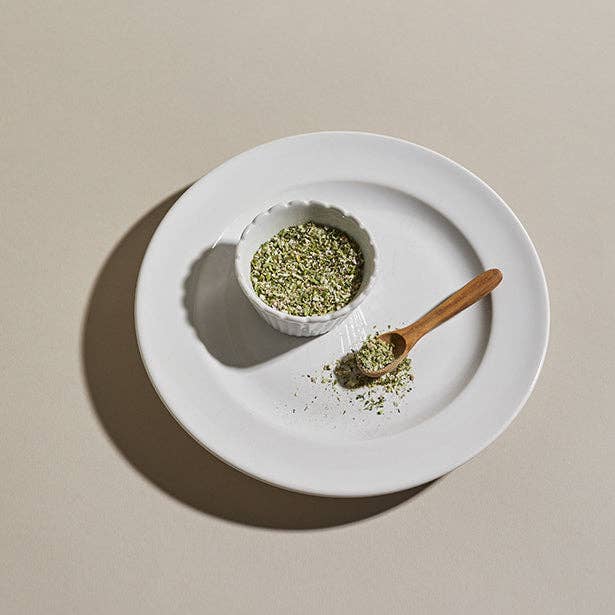 A white scalloped ceramic bowl filled with Parisian Shallot Herb Blend seasoning an a wooden spoon scooping it out onto a plate.