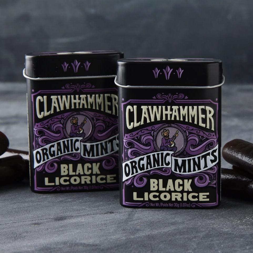 Two black and purple Black Licorice Clawhammer Organic Mints Tins surrounded by pieces of black licorice.