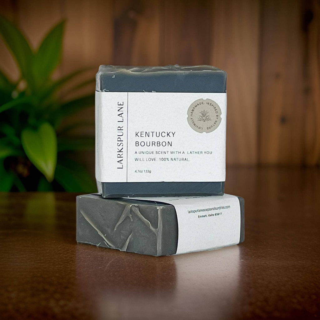 A black bar of Kentucky Bourbon Soap resting on another bar of soap with wood paneled walls and plants behind them.