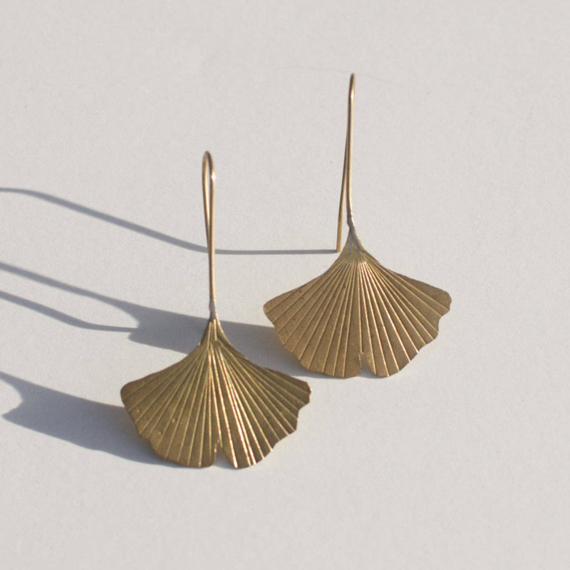 A pair of brass fish hook earrings with a gingko leaf hanging at the end.