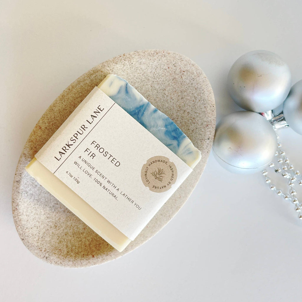 A white and blue swirled bar of Frosted Fir Soap with a cream paper bellyband resting next to a trio of ornaments.