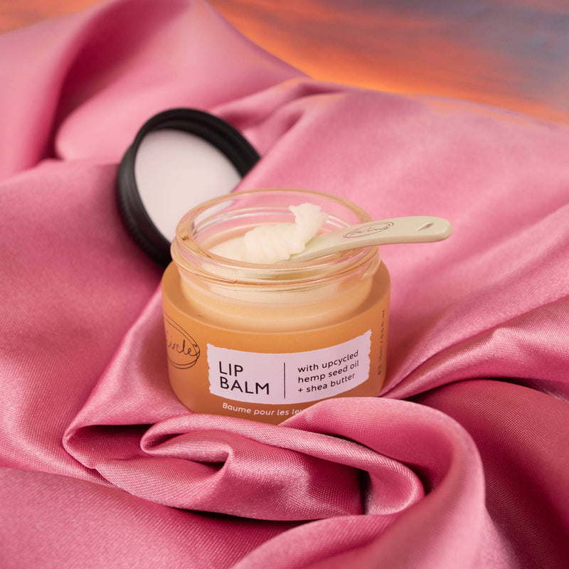 A close-up of an open jar of Lip Balm being spooned out while sitting on a piece of pink fabric.