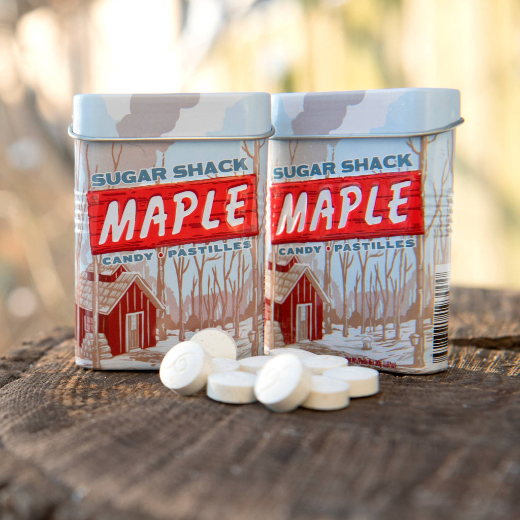 Two Sugar Shack Maple Candy tins ith a pile of maple candies in front on a tree stump.