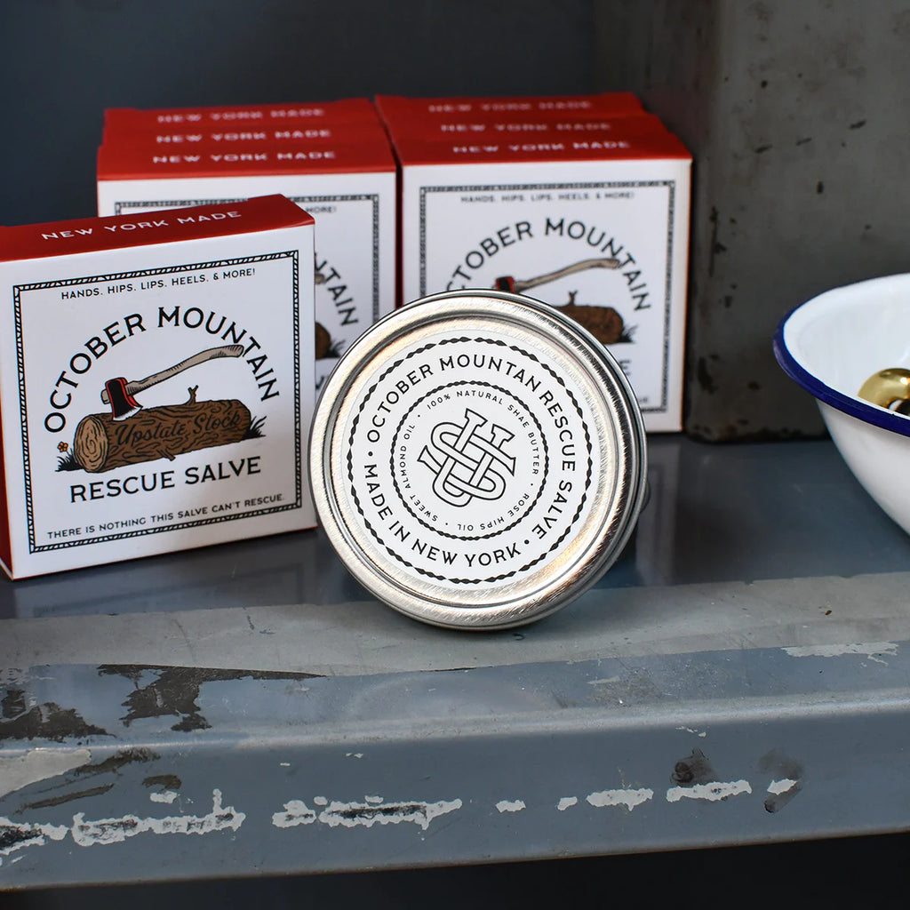 A silver tin of October Mountain Rescue Salve resting on its side in front of two rows of salve boxes.