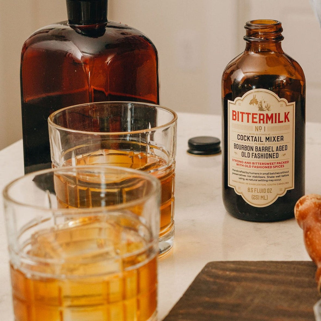 An open bottle of No.1 Bourbon Barrel Aged Old Fashioned next to an amber bottle and two glasses of old fashioned cocktails.