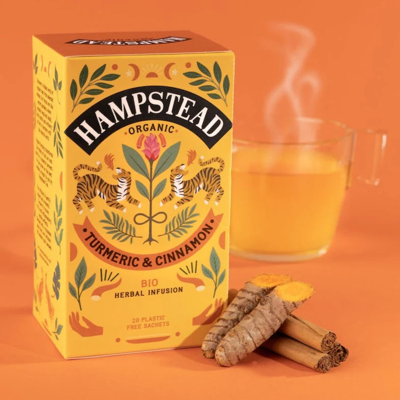 A box of Hampstead Turmeric & Cinnamon Tea next to a pile of ingredients and a glass of freshly brewed tea in a clear glass.