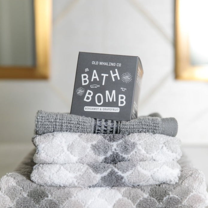 A Bargamot & Grapefruit Bath Bomb box resting on a stack of fluffy gray towels.