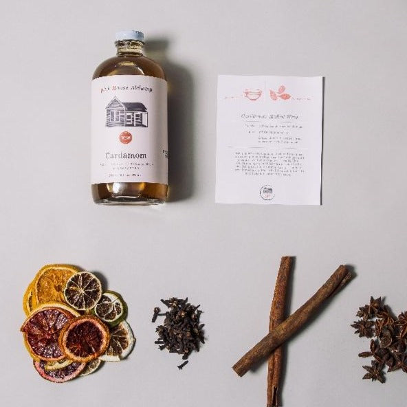 A Cardamom Simple Syrup bottle lying on its side next to a recipe card, dried fruits, star anise, cinnamon, and cloves.